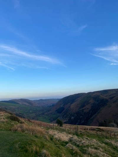 The image presents a landscape scene, likely in the vicinity of Llangynog, United Kingdom. It features a verdant hillside descending sharply into a valley. The grassy terrain is punctuated by a rustic wooden fence running along the slope. A prominent mountain dominates the upper right portion of the image, its steep face showing variations in vegetation. Below the mountain, a valley unfolds with a blend of green fields and forested areas. The sky transitions from a clear blue at the top left to a lighter hue near the horizon, with sparse, thin clouds.