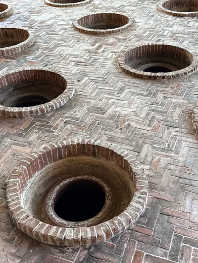 This is a high-angle, close-up, eye-level shot of a brick floor with multiple circular openings. The bricks are laid in a herringbone pattern, with some red and gray variations. The circular openings are lined with bricks and appear to lead into dark voids. Some of the brickwork around the openings shows signs of age and wear. The scene is devoid of people and activity. This appears to be a section of the roof of the Mukhrani Palace in Mukhrani, Georgia, designed for ventilation or light.