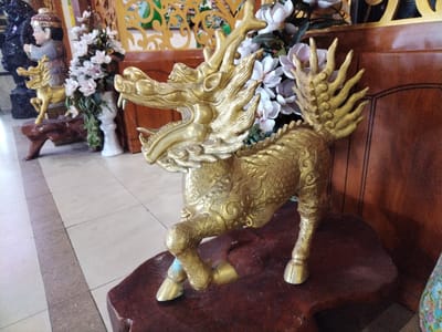 A detailed image captured indoors in Chu Văn An Ward, Vietnam, features a golden, intricately detailed sculpture of a Kirin, a mythical hooved creature, prominently positioned on a dark-stained wooden platform. The Kirin statue has a dragon-like head with an open mouth and flowing mane. The sculpture has a horse-like body covered in intricate patterns and a tail that resembles a fan of feathers.

In the background, elements such as a smaller statue of a child riding a Kirin, a white vase filled with white flowers, and a wooden screen add depth to the composition. A tiled floor in the foreground and a dark wooden panel further back provide texture and visual contrast. The lighting appears to be diffused, highlighting the details of the sculpture and the surrounding objects.