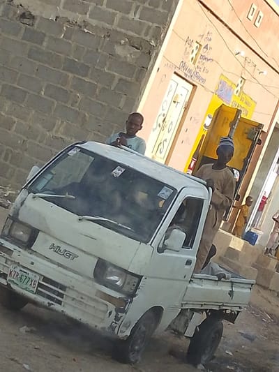 A compact white pickup truck, showing wear and tear, is parked on a street. The license plate reads "KZR-677XB." Two men stand near the truck: one rests his hands on the truck's roof, while the other stands in the truck bed, wearing a traditional cap. In the background, buildings with faded paint and brickwork can be seen, along with hints of other people walking along the street.