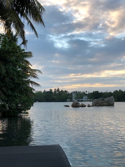 A serene body of water, likely a lake or lagoon, dominates the foreground and midground of the image, reflecting the soft hues of a cloudy sky. In the distance, a dense line of lush green trees forms the shoreline, with palm trees interspersed amongst them. A modern white building with multiple levels is visible behind the trees, hinting at habitation or a resort. Several large, weathered rocks emerge from the water in the midground, adding a natural element to the landscape. On the left side of the frame, the dark, leafy branches of a tree, possibly a palm, overhang the water, creating a natural frame. A dark, flat surface, potentially part of a boat or pier, occupies the bottom left corner of the image, suggesting the vantage point is from the water's edge or a vessel. The sky is filled with a blend of soft grey clouds and patches of pale yellow and orange light, indicating either dawn or dusk. The overall impression is one of tranquility and natural beauty, with hints of human presence. The location is identified as Koggala, Sri Lanka.