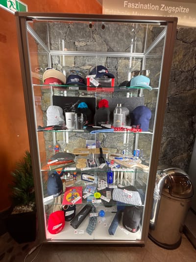 The image displays a glass display cabinet filled with various souvenir items, predominantly hats, sunglasses, and other accessories, likely at the Zugspitze "Erlebnis Museum" in Ehrwald, Austria, as indicated by the text "Faszination Zugspitze Erlebnis Museum" visible at the top of the cabinet. The cabinet is illuminated from within, highlighting the items on multiple shelves. The background consists of a rough stone wall. To the right of the cabinet, a shiny, metallic trash receptacle is visible. The scene is indoors, with a plant in a dark pot to the left of the cabinet. The items in the cabinet appear to be merchandise related to the Zugspitze, an alpine peak.