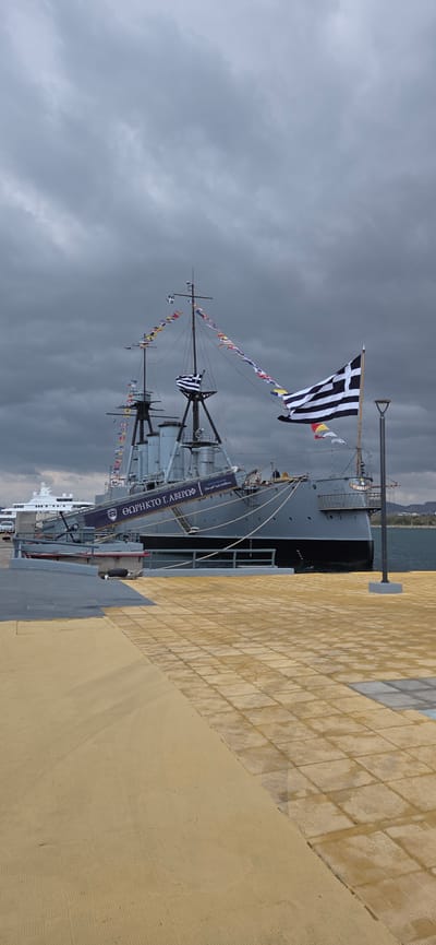 A large, grey naval vessel, identified as the Battleship G. Averof (ΘΩΡΗΚΤΟ Γ. ΑΒΕΡΩΦ) by text on its gangway banner, is prominently docked in a harbor in Palaio Faliro, Greece. The ship features a grey hull above a black waterline, with multiple masts, smokestacks, and a bridge superstructure. Strings of international maritime signal flags are draped between the masts, and a large Greek national flag (blue and white stripes with a cross) flies from a pole at the ship's stern, unfurled to the left. A smaller Greek flag is visible near the bow mast. A gangway, also bearing the ship's name, extends from the vessel to the pier. Indistinct human figures are visible on the ship's deck near the gangway and on the gangway itself.

The foreground is a broad, flat pier constructed from yellow-orange tiles with a repetitive, textured grid pattern. A section of the pier closer to the ship is a darker grey. A tall, grey lamppost stands upright on the pier to the right of the ship's stern. The sky is heavily overcast with dark grey, thick clouds covering the entire upper portion of the frame. The water in the harbor, visible between the ship and the distant land, is calm and appears dark blue-grey. In the background to the left, a white superyacht or large pleasure craft is partially visible, docked further along the pier, with a white vehicle positioned on the dock nearby. Distant landmasses or hills are faintly visible on the horizon to the right, beyond the harbor water. No discernible specific human actions or interactions are clearly depicted.