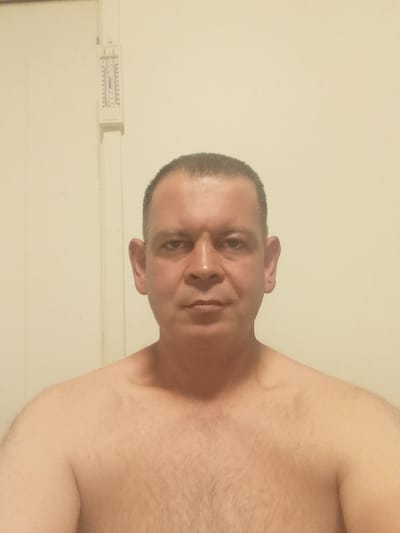 The image shows a medium shot of a man standing shirtless in front of a pale yellow wall. He is facing the camera, with his head and upper torso visible. To the left of the man's head, on the wall, is a vertical white object with markings that resemble a thermometer or ruler. The man has short, dark hair, prominent ears, and a neutral facial expression. The lighting is somewhat dim and appears to come from the front. The background is a plain, slightly textured wall. There is no indication of a city or specific location, including Vallberga, Sweden.