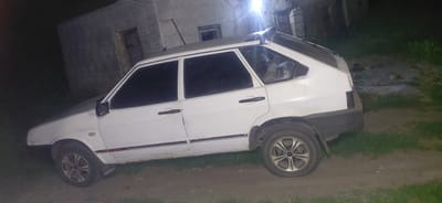 A white hatchback car is parked on a dirt and grass area next to a building at night. The car is positioned at an angle, with its front left wheel and rear right wheel visible. The building in the background appears to be made of cinder blocks and has a weathered appearance. There is a bright light source in the upper right portion of the image, casting a glare. The ground is a mix of dirt and patchy grass. No people are visible in the image. The car has tinted windows and a black roof rack. There are some red accents along the lower side panels of the car. The overall scene suggests a rural or semi-abandoned location in Ketrysanivka, Ukraine, during nighttime.