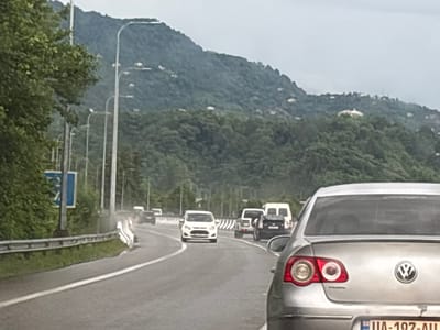 A silver Volkswagen sedan, with license plate UA-197-AU, is in the foreground, facing away from the camera. The car is on a highway with multiple lanes, indicated by white lane markings. In the background, there are several other vehicles, including a white van and a dark SUV. The highway is bordered by a metal guardrail and lush green vegetation. Beyond the highway, a steep, tree-covered mountainside rises, with scattered white buildings visible on its slopes. The sky is overcast, suggesting a cloudy day. The scene appears to be located on a highway near Kveda Kvirike, Georgia, given the context.