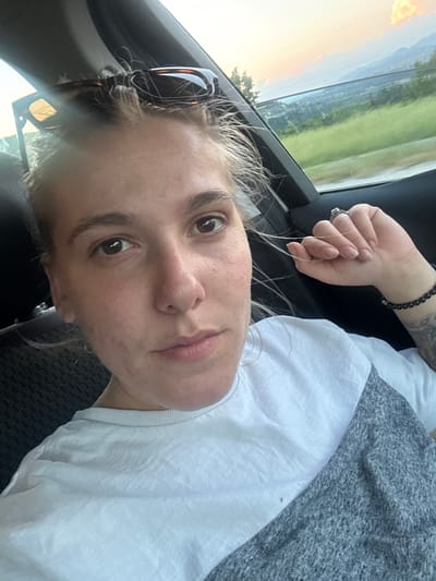 A light-skinned individual is depicted in a close-up, chest-up selfie taken inside a vehicle, located in Dobrotino, Bulgaria. The person, appearing to be female, looks directly into the camera with a neutral expression. She has light-colored hair, some strands loose around her face, and a pair of tortoiseshell-patterned sunglasses resting atop her head. She is wearing a two-tone t-shirt, white on the upper portion and heather grey on the lower-right side. Her left hand is raised, with the index finger and thumb casually holding a strand of her hair; a silver-colored ring with a stone is visible on her left ring finger. A dark beaded bracelet adorns her right wrist, and a portion of a dark tattoo is visible on her right forearm. The car's interior features dark upholstery and trim. Through the vehicle window, a landscape is visible under a soft-hued sky (pinks, oranges, light blues), indicating either sunrise or sunset. The background includes green fields, distant hills or mountains, and faint structures in the midground.