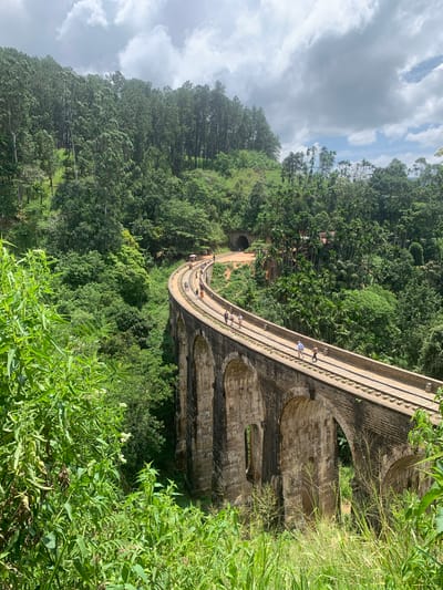 A railway bridge with nine arches, constructed from stone and brick, curves through a lush, green landscape. The bridge is elevated above a forested valley, with dense trees and vegetation on either side. Several people are walking along the train tracks on top of the bridge, some in small groups and others individually. In the background, a tunnel entrance is visible on the left, and scattered buildings can be seen amidst the trees on the right. The sky above is partly cloudy. This is the Nine Arch Bridge in Ella, Sri Lanka.