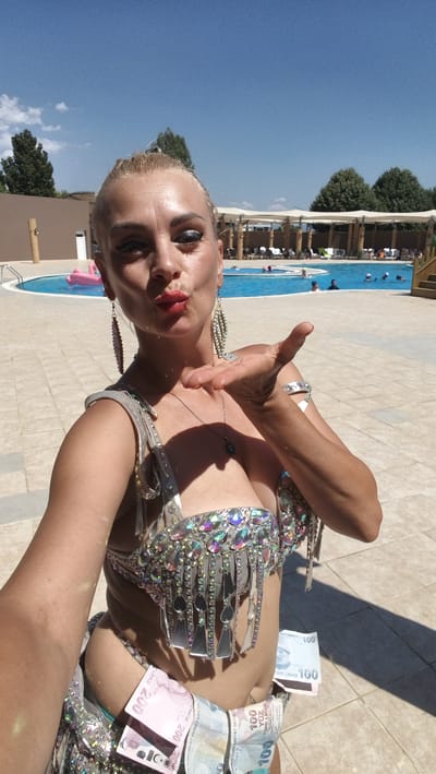 A woman, possibly in Ödemiş, Turkey, poses for a selfie by a swimming pool. She is wearing a bejeweled costume and has money tucked into her waistband. The woman is blowing a kiss towards the camera. The pool area is paved with light-colored tiles and features lounge chairs and umbrellas. Trees and a building are visible in the background, along with a pink inflatable object in the pool. The sky is clear and blue.