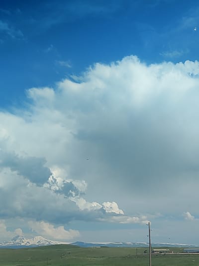 A wide, eye-level, full shot captures a vast expanse of sky over a rural landscape in Azatan, Armenia. The sky is predominantly a vibrant blue, with dramatic, puffy white and gray clouds dominating the upper half of the frame. In the far distance, a majestic mountain range, capped with snow, stretches horizontally across the horizon. The foreground consists of rolling green hills, dotted with a few sparse trees and a single, tall wooden utility pole. A small, dark airplane is visible in the sky, flying towards the left side of the frame. The overall scene suggests a bright, possibly windy, day with a sense of open space and natural beauty.