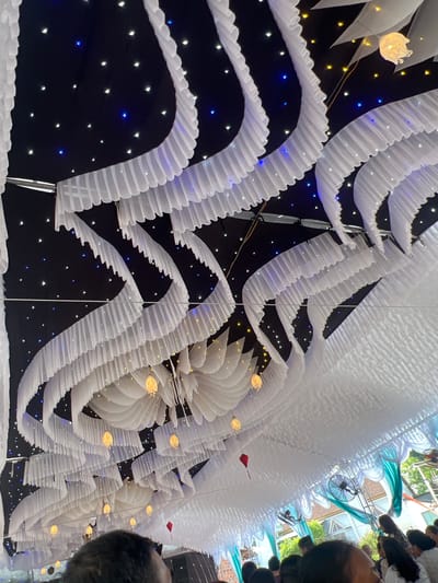 The image shows a street decoration in Bình An, Vietnam. The scene is an under-looking view of a street covered with overhead decorations. These consist of white ruffled streamers arranged in arches, spanning the street. The background above the streamers is black, dotted with twinkling blue and white lights, giving the impression of a night sky. Interspersed among the streamers are white flower-like structures, also illuminated. A few red flags are also visible among the white decorations. In the upper right of the image, people can be seen walking along the street, along with a bicycle. The light indicates it is daytime despite the decorations.