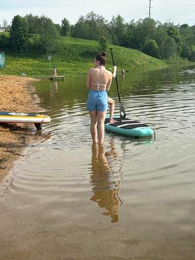 A woman in a white bikini top and blue shorts is lying prone on a paddleboard in a lake. She is holding a paddle. Another paddleboard rests on the sandy shore in the foreground. The lake is bordered by a grassy shoreline with trees in the background. The location is Novosinkovo, Russia.