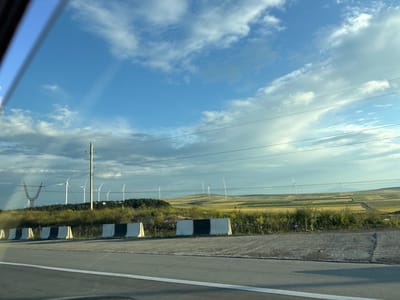 The image depicts a view of a rural landscape with wind turbines, taken from a moving vehicle in the vicinity of Urbnisi, Georgia. The foreground shows a paved road with a white line and concrete barriers with black and white markings. Beyond the road, there is a grassy embankment dotted with some litter. In the middle ground, a line of wind turbines stands against a backdrop of rolling hills. The hills are divided into fields of various colors, suggesting agricultural activity. Power lines are visible crisscrossing the sky and connecting to a tall pylon on the left. The sky is partly cloudy with a mix of blue sky and white and gray clouds. The overall scene suggests a bright, possibly late afternoon or early morning lighting condition. No people are visible in the image.
