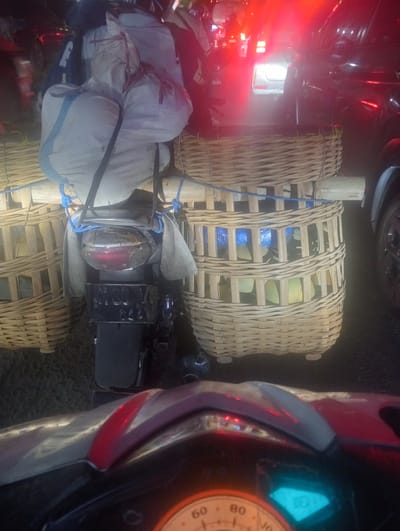 The image shows a motorcycle heavily laden with goods, possibly for transport or sale. The motorcycle is seen from behind, with its taillight and license plate partially visible. Large woven baskets are strapped to the back of the motorcycle, containing what appear to be produce, possibly fruits or vegetables, wrapped in plastic. The load is secured with ropes.
The scene appears to be a busy street in Sruni, Indonesia, likely at night given the artificial lighting and the presence of vehicle headlights and taillights in the background. The red glow from vehicle lights suggests traffic and a lively atmosphere. The motorcycle's dashboard, with a speedometer dial, is visible in the foreground, indicating the perspective of someone riding or observing from the motorcycle.
The overall impression is one of commerce and daily life in an Indonesian setting, with a focus on the practicalities of transporting goods by motorcycle. The time of day is likely evening or night, and the weather appears to be clear, allowing for visibility of the traffic. The mood is functional and perhaps a bit chaotic, typical of busy traffic conditions. No discernible text is clearly readable in the image, apart from markings on the motorcycle's speedometer.