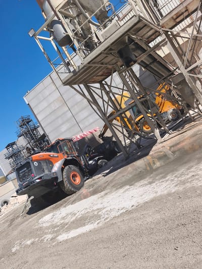 In Nàquera, Spain, an outdoor industrial scene unfolds under a clear blue sky. The image captures a large orange wheel loader, identified as a DL380, with the brand name "MAQUINZA" visible, positioned on a dusty, gravelly surface. The loader has a speed limit sign of "40" displayed prominently. To the right of the wheel loader, a yellow backhoe loader is partially visible, its tires and front loader arm suggesting it is also at work. Dominating the background is a tall, multi-level industrial structure, likely part of a cement or aggregate processing plant, constructed with metal beams and platforms, and featuring large cylindrical silos. A light gray, rectangular building stands to the left of the industrial structure. The ground is a mix of dry gravel and damp, muddy patches, indicating recent activity or a spill. The overall impression is one of an active construction or material processing site.