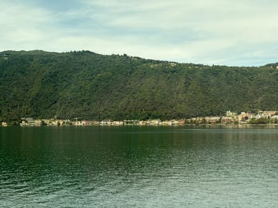 This is a horizontal, medium-shot, eye-level, outdoor, daytime photograph of the lakefront of Melide, Switzerland. The foreground is dominated by a wide expanse of dark green, choppy water, with distinct ripples reflecting the light. In the midground, a densely forested, green mountain rises steeply from the water's edge. Along the base of the mountain, a line of buildings forms the town of Melide. These buildings are varied in color and architectural style, ranging from light yellow and white to reddish-brown. Some appear to be apartment complexes or hotels, with multiple stories and balconies. A few scattered trees are visible between the buildings. Towards the right side of the midground, a yellow construction crane is visible above the rooftops. The background consists of a hazy, pale blue sky with thin, wispy clouds. The overall impression is of a scenic lakeside town nestled against a forested mountainside.