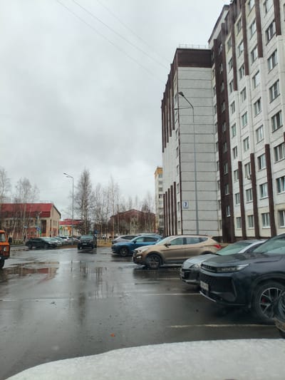 The image depicts a street scene in Kogalym, Russia on an overcast day. The street is wet, suggesting recent rain or snow melt, and there is some snow visible near the bottom of the frame. Parked cars line the street, including a brown Hyundai Santa Fe and a black car in the foreground with the license plate "E380XH 186". In the background, a multi-story apartment building with brown and white exterior walls dominates the right side of the frame. A street sign with the number "7" is visible on the side of the building. Across the street, there is a lower, red-roofed building and leafless trees. A traffic light is visible at a street intersection.