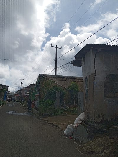 This daytime photograph depicts a narrow, paved road in a residential area of Hinabangan, Philippines, under a partly cloudy sky with bright sunlight. Numerous utility lines crisscross overhead, strung between several power poles that stretch down the street.

On the right side of the foreground, a dilapidated concrete building with a corrugated metal roof is prominent. It features a significant vertical crack running down its facade, indicating disrepair. Two white sacks rest at its base along the roadside. Further along the road, additional modest residential structures are visible, some partially obscured by overgrown vegetation.

On the left side of the road, two figures, likely women, are standing near a small stall or structure. One individual appears to be wearing a head covering. They are somewhat obscured by the distance. The road itself shows several wet patches or puddles. The scene suggests a local street in a rural or semi-urban setting, characterized by weathered buildings and visible utility infrastructure. No discernible text is visible within the image.