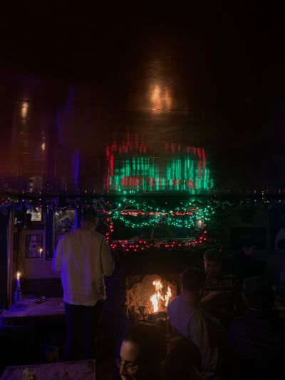 A mirrored room showcases festive lighting with a fireplace scene. Several individuals gather around the fireplace while one person stands in front of a bar counter, illuminated by blue light. The mirror reflection doubles the array of red and green Christmas lights, enhancing the overall festive ambiance.
