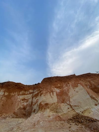 The scene depicts a large, exposed earth cliff or embankment in Asaga Amangwu, Nigeria, dominated by stratified soil layers under a partially cloudy sky. The upper portion of the image, roughly 40-50%, shows a light blue sky with a distinct vertical division: the left side presents a clearer, brighter blue with wispy white clouds, while the right side features denser, lighter gray and white clouds. The lower 50-60% of the image consists of a weathered earth face. The uppermost layer of the cliff is a rich, reddish-brown soil, likely lateritic, with minimal, sparse dry vegetation visible along its crest. Below this, the cliff face reveals prominent, irregularly shaped horizontal and diagonal bands of a lighter, creamy white or pale yellow material, which appears to be clay or sand, intermixed with streaky reddish-brown deposits. The textures suggest erosion and fracturing. At the immediate base of the cliff, especially towards the right, piles of loose reddish-brown soil and fragmented rocks are visible, indicating recent material collapse or excavation. No people or animals are present.
