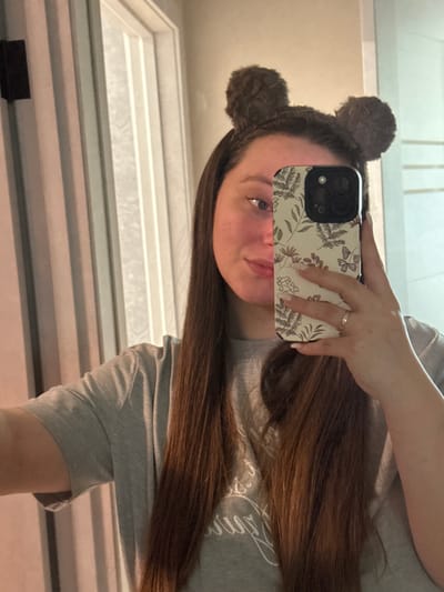 A forensic examination of the image reveals a mirror selfie taken indoors within the city of Tliustenkhabl', Russia. A female individual, characterized by long, straight brown hair and fair skin, is depicted. She wears a grey short-sleeved t-shirt and a dark brown fluffy headband fashioned with two pom-poms resembling animal ears. Her right arm is extended, holding a smartphone with a botanical-patterned case (featuring leaves, flowers, and butterflies in brown tones) and a multi-lens camera array, which partially obscures the left side of her face. Her visible right eye is blue, and she exhibits a slight smile. A gold-toned ring is present on the ring finger of her right hand, which also displays light-colored manicured nails. The background, visible through the mirror's reflection, consists of an interior space featuring a light-colored door with vertical paneling.