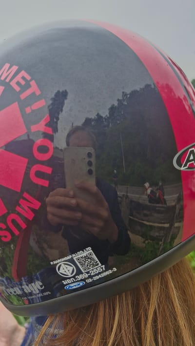 The image is a reflection on a motorcycle helmet in Samoeng Tai, Thailand. A person is holding a smartphone and taking a selfie, with their face and hands partially visible. The helmet has red and black graphics, including lettering that reads "MET! YOU MUS". A sticker with a QR code and Thai text is on the helmet. In the reflection, a rural road with trees, a stone wall, and a few motorcycles are visible. The sky is overcast.