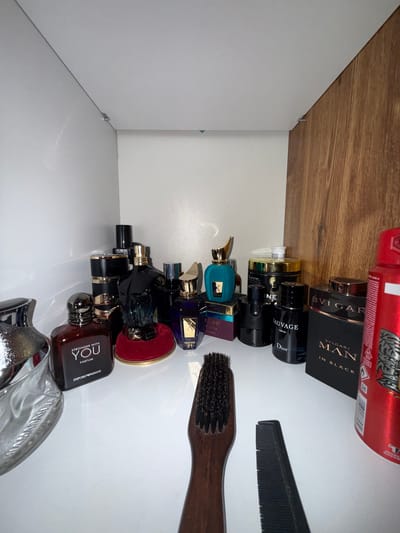 A collection of men's fragrances is displayed on a white shelf against a white wall with a wooden panel on the right. From left to right, a partially visible clear glass bottle with a silver cap and embossed text is present. Next to it is a square, dark red bottle labeled "STRONGER WITH YOU PARFUM EMPORIO ARMANI." To the right of this is a dark blue, muscular-shaped bottle resting on a red velvet display stand. Behind this, a tall, dark bottle with a gold cap is visible. Further to the right, a teal-colored bottle with a gold stopper is placed on a purple and gold box. Next to it is a black bottle labeled "SAUVAGE DIOR." Behind that is a black jar with a gold lid and some white text, followed by a black bottle with a silver cap. To the right of these is a black box labeled "BVLGARI MAN IN BLACK." On the far right, a tall, red cylindrical object, possibly an aerosol can, with silver and black detailing is positioned. In the foreground, a dark wooden-handled brush and a black comb lie on the shelf. The scene appears to be an organized display of personal grooming products. The location is a shelf in a room, with no visible indicators of the city of Brenitsa, Bulgaria.
