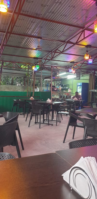 This image depicts the interior of an open-air restaurant. The structure has a corrugated metal roof supported by red metal beams. Colorful lights, casting a rainbow effect on the ceiling, are suspended from the roof, along with a ceiling fan. The floor is a concrete patio. Dark-colored tables with matching chairs are arranged throughout the space. In the background, a man in a white shirt is visible near a green corrugated metal wall. Further back, another man is seated at a table. To the right, there's a blue refrigerator and a counter area with various items. The restaurant appears to be located in Chor Sahapur, Bangladesh.