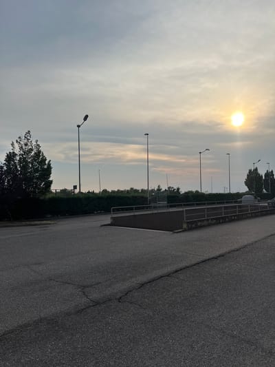 The image depicts an outdoor scene at what appears to be a parking lot or an industrial area during twilight. The sky is partly cloudy with a bright sun peeking through the clouds in the upper right quadrant, casting a soft glow over the scene. Several tall streetlights are visible, dotting the landscape. In the foreground, a cracked asphalt surface dominates the lower portion of the image. A concrete ramp with metal railings leads upwards from left to right. Beyond the ramp, a dense hedge forms a barrier, behind which several trees are silhouetted against the sky. In the distance, more streetlights and some structures are faintly visible. A single vehicle, possibly a trailer, is parked on the right side. There are no people visible in the image. The overall impression is one of a quiet, utilitarian environment at the end of the day. The location is San Martino Buon Albergo, Italy.