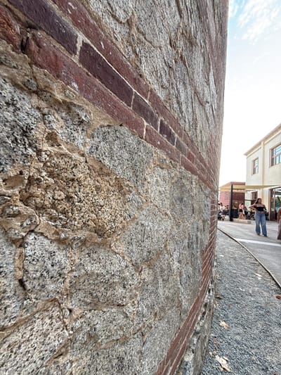 The image depicts a close-up view of an exterior brick and stone wall in Mataró, Spain. The wall is weathered, with sections of rough-hewn gray stone interspersed with red brick. The stone exhibits a coarse texture with visible aggregate, while the bricks are arranged in a horizontal pattern. In the background, a pathway or street is visible, lined with gravel and a metal track, possibly remnants of a railway. Beyond the pathway, buildings with light-colored facades and windows are present. Several individuals are scattered in the background, some walking and others seated at outdoor tables, suggesting a public or commercial area. The sky above is bright with scattered clouds.