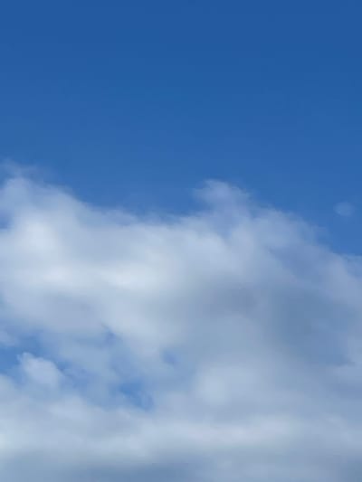 The image depicts a bright blue sky with scattered, soft white clouds. The clouds are primarily located in the lower half of the frame, appearing somewhat wispy and diffused, suggesting either a sunny day with light cloud cover or possibly a hazy atmosphere. The upper portion of the image is dominated by uninterrupted blue sky. There are no visible structures, people, or identifiable landmarks within the frame. The lighting suggests it is daytime. The context of Sviatopetrivske, Ukraine, implies this is an outdoor scene, likely a view looking upwards.