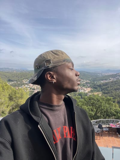 A young Black male wearing a dark grey t-shirt with red text and a black zip-up hoodie is pictured from the chest up, facing right with his eyes closed. He is wearing a camouflage baseball cap. The background shows a landscape of rolling green hills dotted with houses, indicating an urban area within a natural setting. The location is identified as Can Güell, Spain. A tiled patio with tables and chairs is visible in the lower right corner. The sky above is a light blue with scattered clouds.