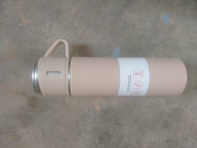 A light pink cylindrical thermos bottle lies horizontally on a concrete surface. The bottle has a silver cap on the left and a light pink handle extending upwards from the cap area. A white label with red text and icons is wrapped around the middle of the bottle. The concrete surface is textured and appears to be outdoors. The scene is static, with no people or visible actions taking place. The lighting is natural and appears to be daylight. The location of Majemu, Nigeria is not discernible from the image itself.