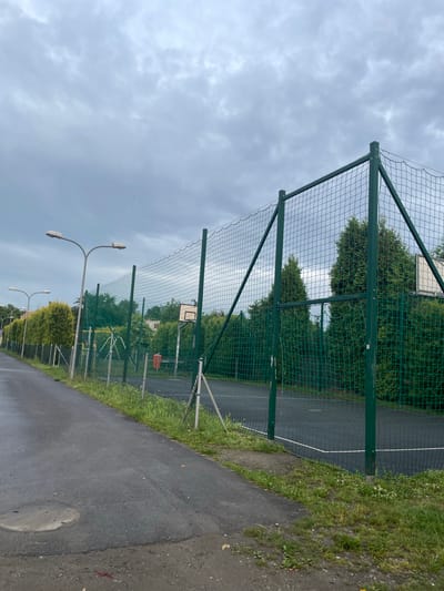 The image captures an outdoor basketball court in Wysoka, Poland, under an overcast sky. A dark asphalt path borders the left side of the frame, with a green chain-link fence enclosing the court. The court itself has a dark surface with white boundary lines. A basketball hoop with a backboard is visible inside the fenced area, positioned amidst several tall, dense evergreen trees that surround the court. Streetlights line the path, and a small orange trash receptacle is also present near the fence. The ground is covered in a mixture of grass and patchy dirt, with some debris visible on the asphalt.