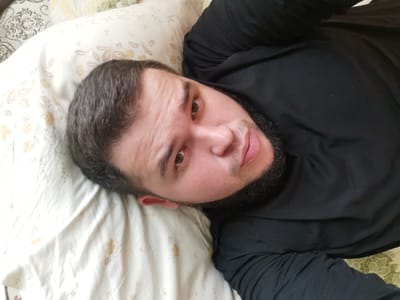 The media file is a selfie taken by a man lying on a bed. He has dark hair and a beard and is wearing a black long-sleeved shirt. He is looking directly at the camera with a neutral expression. The background consists of a pillow with a floral pattern and a blanket. The lighting suggests it is daytime, and the indoor setting of a bedroom is implied by the bed. No text is visible in the image. The location context is Serov, Russia.
