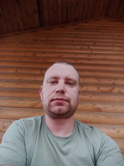 This is a portrait of a man, taken from a close-up, slightly upward angle, with his head tilted. He has short, light brown hair and stubble on his chin and upper lip. His eyes are open and looking towards the camera, and his lips are slightly parted. He is wearing a light green t-shirt.

The background consists of wooden paneling. The wood appears to be pine or a similar softwood, with visible grain and knots. The lighting suggests an indoor setting, possibly a rustic cabin or a room with wooden walls. The overall mood is casual and informal. There is no visible text in the image. The location is identified as Падгорʼеўскі сельскі Савет, Belarus, which is a rural administrative unit in Belarus.