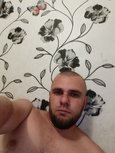 A close-up, selfie-style, chest-up image features a light-skinned, bald man with a beard looking directly at the camera. He is topless, revealing his left arm and shoulder. The background consists of a white wall with a repeating pattern of black and white flowers and a single colorful butterfly. The setting appears to be indoors. The image was taken in Pishchane, Ukraine.