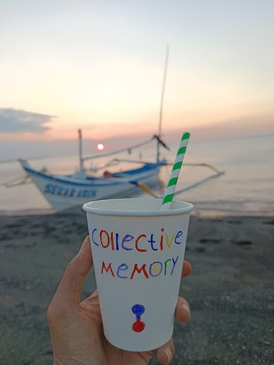 A close-up shot features a hand holding a white paper cup with colorful writing on it. The cup has a green and white striped straw peeking out of the top. The writing on the cup spells out "COLLECTIVE MEMORY" in various colors. Below the text, there is a small drawing of what appears to be a rattle, also in red and blue.
In the background, a serene beach scene unfolds. A traditional Indonesian fishing boat, with outriggers and a sail, is anchored near the shore. The boat has text on its side that reads "SEKAU ARUN." The sun is setting over the ocean, casting a warm, soft glow across the sky, with hues of orange and pink visible. The water is calm, and the shoreline is covered in dark sand or pebbles. The overall atmosphere is peaceful and evokes a sense of nostalgia or reflection, enhanced by the sunset and the message on the cup. The time of day is clearly sunset. The weather appears to be calm and pleasant, suitable for a beach setting.