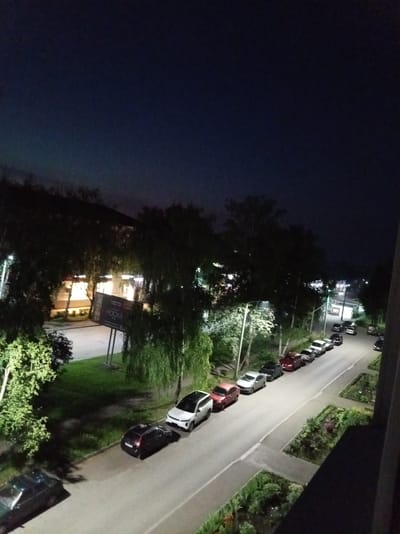 The image shows a nighttime scene in the city of Alatyr, Russia. A street is lined with parked cars, mostly sedans and hatchbacks, illuminated by streetlights. Trees with dense foliage border the street and a sidewalk. In the background, a building with brightly lit windows is visible, and a sign for "HOOKA" is mounted on a stand near the building. The sky above is dark blue, indicating it is nighttime. The overall impression is of a quiet urban environment at night.