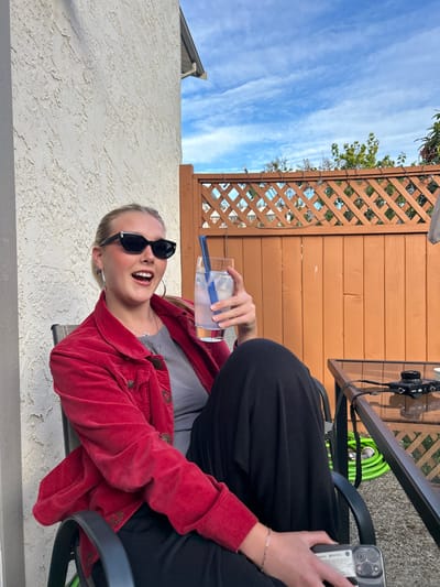 A young female is seated outdoors in a residential backyard or patio setting, situated in Victoria, Canada. She is positioned facing slightly right of the camera, wearing black cat-eye sunglasses, large silver hoop earrings, a red corduroy jacket over a grey t-shirt, and black wide-leg pants. Her blonde hair is pulled back, and she is smiling broadly with her mouth open. Her left hand holds a clear glass containing a light purple liquid, ice cubes, and a blue reusable straw. Her right hand rests on her lap, holding a grey smartphone. To her right, a black compact camera with a lens is visible on a dark-framed glass-top patio table. The background features a light-colored stucco building wall on the left, a tall brown wooden fence with a lattice top section, and green foliage visible above the fence line. A white rain gutter runs along the top of the stucco wall. A green coiled garden hose is partially visible on the ground behind the table. The sky above is blue with scattered white clouds.