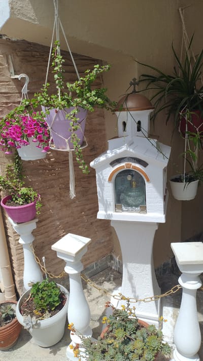 This image depicts an outdoor scene in Νέα Μηχανιώνα, Greece. The central focus is a small, white, church-like structure mounted on a pedestal. This structure has a domed roof with a cross on top and a glass-fronted opening displaying religious iconography, likely a saint or an icon, along with a glass oil lamp. Around the base of this structure, and on the ground, are various potted plants. These include succulents in terra cotta and white pots, and a flowering plant with pink blossoms hanging in a white pot suspended by macrame. Another hanging basket with a trailing green plant in a purple pot is also visible. The background features a textured, light brown brick wall and a pale, possibly stuccoed, overhang. White balusters connected by a gold chain are positioned in the foreground, suggesting a pathway or a decorative border. The lighting indicates it is daytime, with distinct shadows present. There are no people visible in the scene.