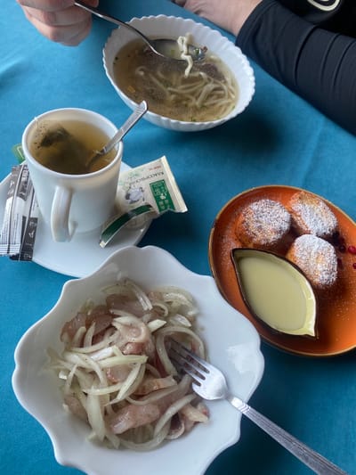 The image captures a meal consisting of soup with noodles in a white bowl, herring with sliced onions in another white bowl with a fork stuck in it, and small round cakes dusted with powdered sugar served with a dipping sauce on an orange plate. A cup of tea with a tea bag and sugar packets accompanies the food. The table is covered with a blue cloth. A person's arm is visible on the left side of the image.