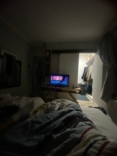 The image shows a bedroom in a house located at latitude -26.139 and longitude 28.352 in the city of Benoni, South Africa. The room is dimly lit, with a bed in the foreground, a television on a table in the middle ground, and a closet in the background. The bed has a white blanket and a blue and white striped comforter on top. The television is on and playing a show. There is a book on the bed and a pair of white socks on the floor. The closet is full of clothes. The room is in a state of disarray, with items scattered about. There are no people in the image. The environment is quiet and serene.