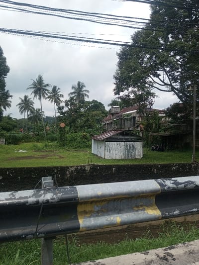 This image captures an outdoor scene in Chori, Nigeria, under an overcast sky. In the foreground, a weathered metal guardrail, painted gray and black with significant peeling yellow and black paint, runs horizontally. Below the guardrail, a concrete wall exhibits dark discoloration and moss, with green grass visible at its base.
The midground features an expansive, unkempt grassy field. A small, rectangular, single-story structure with corrugated metal siding and an exposed roof frame stands on the right side of this field. To its right, a larger, multi-story building with a reddish-brown tiled roof appears structurally compromised or incomplete, partially obscured by dense green foliage. Two dark-colored motorcycles are parked near this larger building. A utility pole with a circular, dish-like object is visible in the left-midground, amidst overgrown vegetation. Another tall light pole stands on the far right.
The background is characterized by numerous tall palm trees and dense, dark green tropical foliage extending across the horizon. A distant, green-covered hillside is visible on the left. The sky is uniformly gray with heavy cloud cover. Multiple dark overhead power lines traverse the upper portion of the frame. No individuals, animals, or active subjects are present.