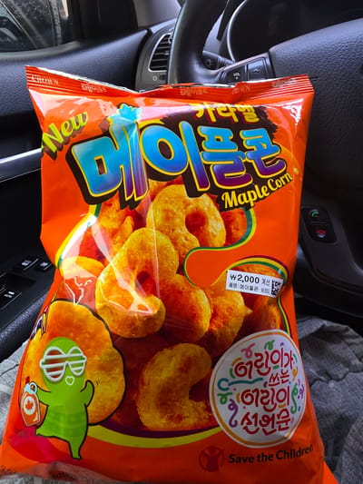 The image shows a bag of "Maple Corn" snack food placed on a car seat. The bag is primarily orange with colorful text and graphics. A prominent graphic of a green cartoon character holding a maple syrup bottle is on the left side of the bag. In the upper right corner of the bag, there is a circular logo with Korean text and the phrase "Save the Children" in English. The car's steering wheel and dashboard are visible in the background, indicating the setting is inside a vehicle. The lighting suggests it is daytime. There are no people visible in the image. The car appears to be parked, likely in a location within Daejeon, South Korea, given the Korean text on the packaging.