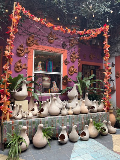 A building facade decorated with terracotta-colored carvings of animals and draped with autumn-leaf garlands. A window displays colorful textiles and pottery. A table exhibits a collection of light-colored, stylized animal sculptures, some with plants growing inside. The sculptures are arranged in rows and appear to be made of clay or ceramic. The setting suggests a crafts store or artisan's workshop.