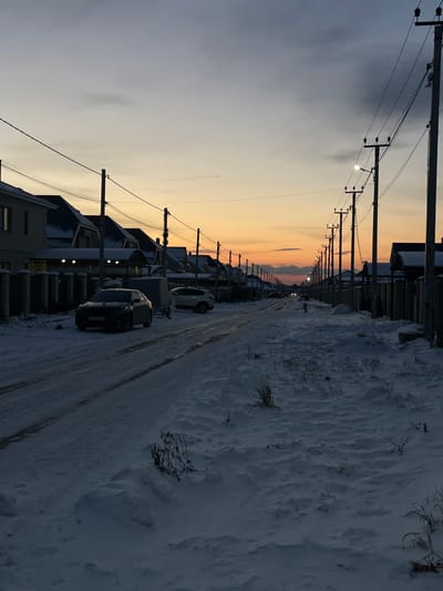 The image depicts a snow-covered residential street in Tsibanobalka, Russia, at dusk or dawn. The sky exhibits a gradient from muted grey-blue overhead to prominent orange and yellow hues at the horizon. A straight, unpaved road, covered in compacted snow and ice with visible tire tracks, recedes into the distance, flanked by deep snow on both sides.

On the left side of the street, a dark-colored sedan, possibly a Toyota Camry, is parked facing the viewer, partially on the snowy shoulder. Behind it, a small enclosed utility trailer is present. Further down the street on the left, a light-colored SUV is also parked. No occupants are visible in or around the vehicles.

Numerous single-family houses, primarily two-story structures with dark roofs mostly covered in snow, line both sides of the street. Fences of varying styles separate the properties from the road. Some houses exhibit active exterior lighting fixtures.

A continuous line of utility poles with crossarms and overhead wires extends along both sides of the street, converging in the distance. Several pole-mounted streetlights are illuminated, casting artificial light onto the scene. No individuals are present in the observable area. The environment is consistent with a cold, clear winter twilight.