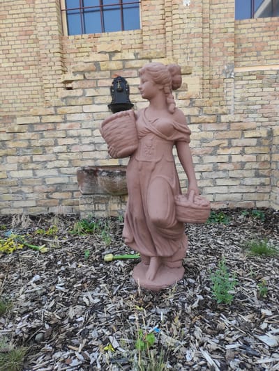 The image displays a reddish-brown statue of a woman carrying two baskets, one on her left arm and the other in her right hand. The statue is situated in front of a light tan brick building with a visible window pane and a dark fixture mounted on the wall above and behind the statue, possibly a water spigot. The ground in front of the building is covered with dark wood chips and some scattered greenery and fallen yellow flowers. The statue is positioned on a base and is the central focus of the foreground. The background consists of the brick wall of the building and the ground covering. The overall impression is an outdoor decorative setting.