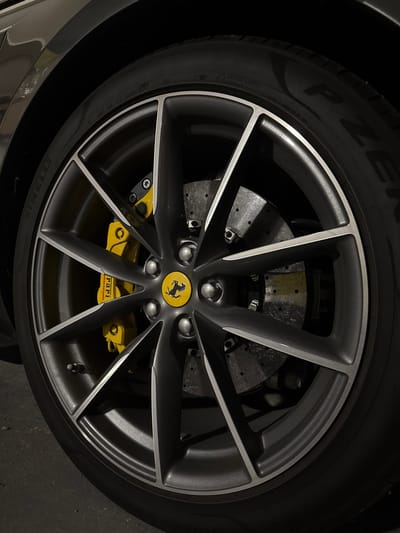 The image displays a detailed close-up of the front-right wheel and tire assembly of a dark-colored Ferrari, located in Lège-Cap-Ferret, France. The vehicle is equipped with multi-spoke alloy wheels, finished in a matte dark grey with polished silver accents on the edges of each spoke. A yellow center cap, featuring the black Ferrari prancing horse emblem, is precisely positioned. Behind the wheel spokes, a bright yellow brake caliper is clearly visible, bearing the "Ferrari" inscription and indicating "carbon ceramic" brake technology. A large, perforated carbon-ceramic brake rotor is partially visible. The tire is a Pirelli P Zero, with elements of its branding and manufacturing codes visible on the sidewall. The setting appears to be an indoor or dimly lit environment, with subtle reflections on the car's body panel in the upper left quadrant. The ground beneath the tire is a dark, textured surface consistent with concrete or asphalt. No individuals are present in the scene.