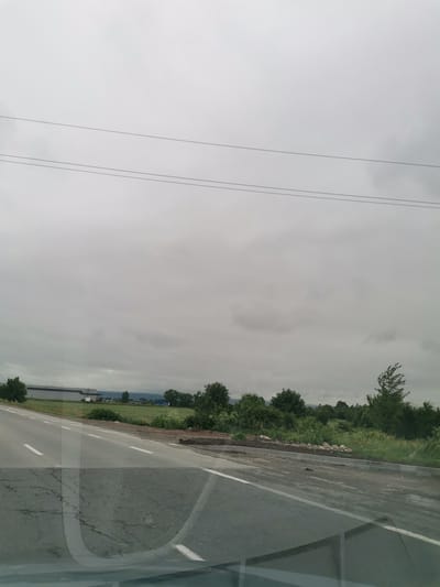 The image shows a roadside view on a cloudy day in Krivina, Bulgaria. A paved road with white lane markings runs diagonally from the bottom left to the middle of the frame. The road appears to be under construction or repair on the right side, with loose stones and dirt visible. Beyond the road, a grassy field stretches to a line of trees. In the distance, a large industrial building with a light-colored facade is visible on the left. A few more buildings and trees are scattered across the landscape, with mountains visible in the far distance. Overhead, power lines stretch horizontally across the sky. The overall scene depicts a rural or semi-industrial area with overcast weather conditions.